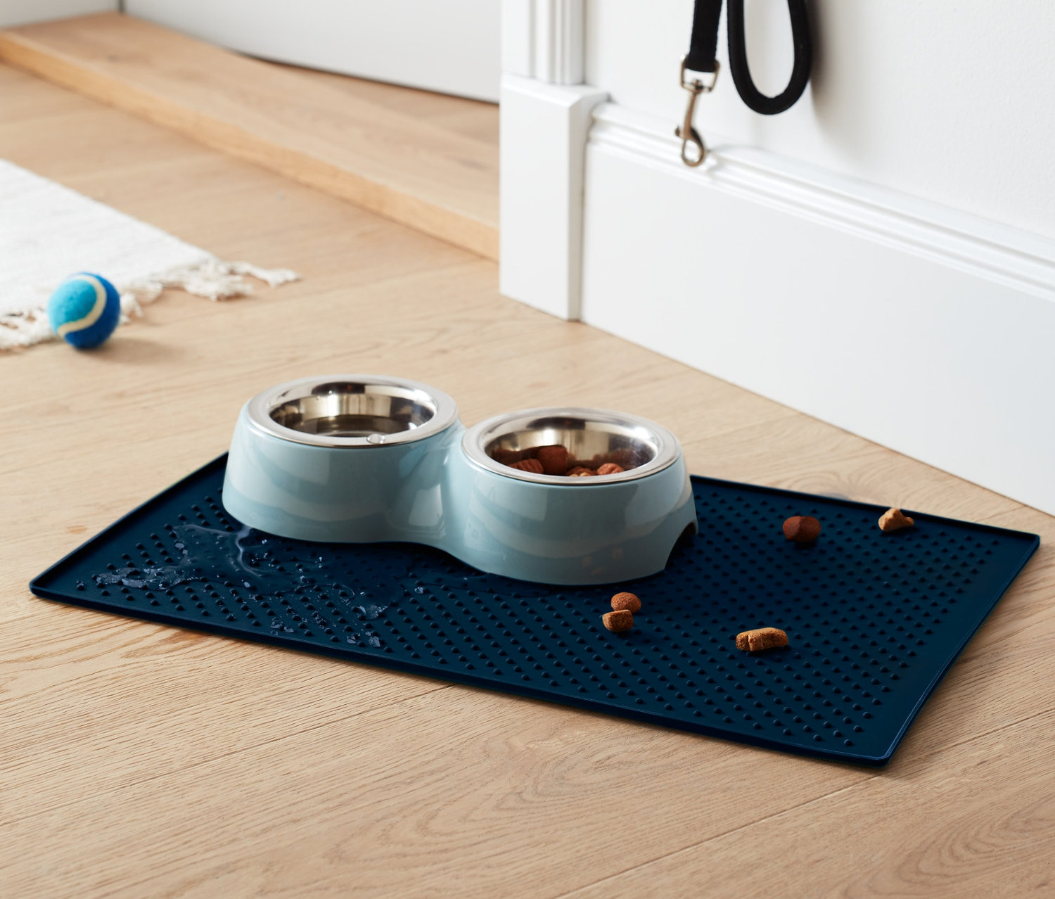 Niebieska podkładka pod miskę dla zwierząt z dwiema miskami ze stali nierdzewnej, jedna z jedzeniem, leży na drewnianej podłodze.