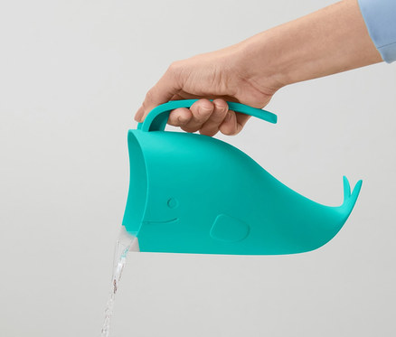Ręka trzyma turkusowy kubek do mycia włosów dla dzieci w kształcie wieloryba, z którego wylewa się woda.