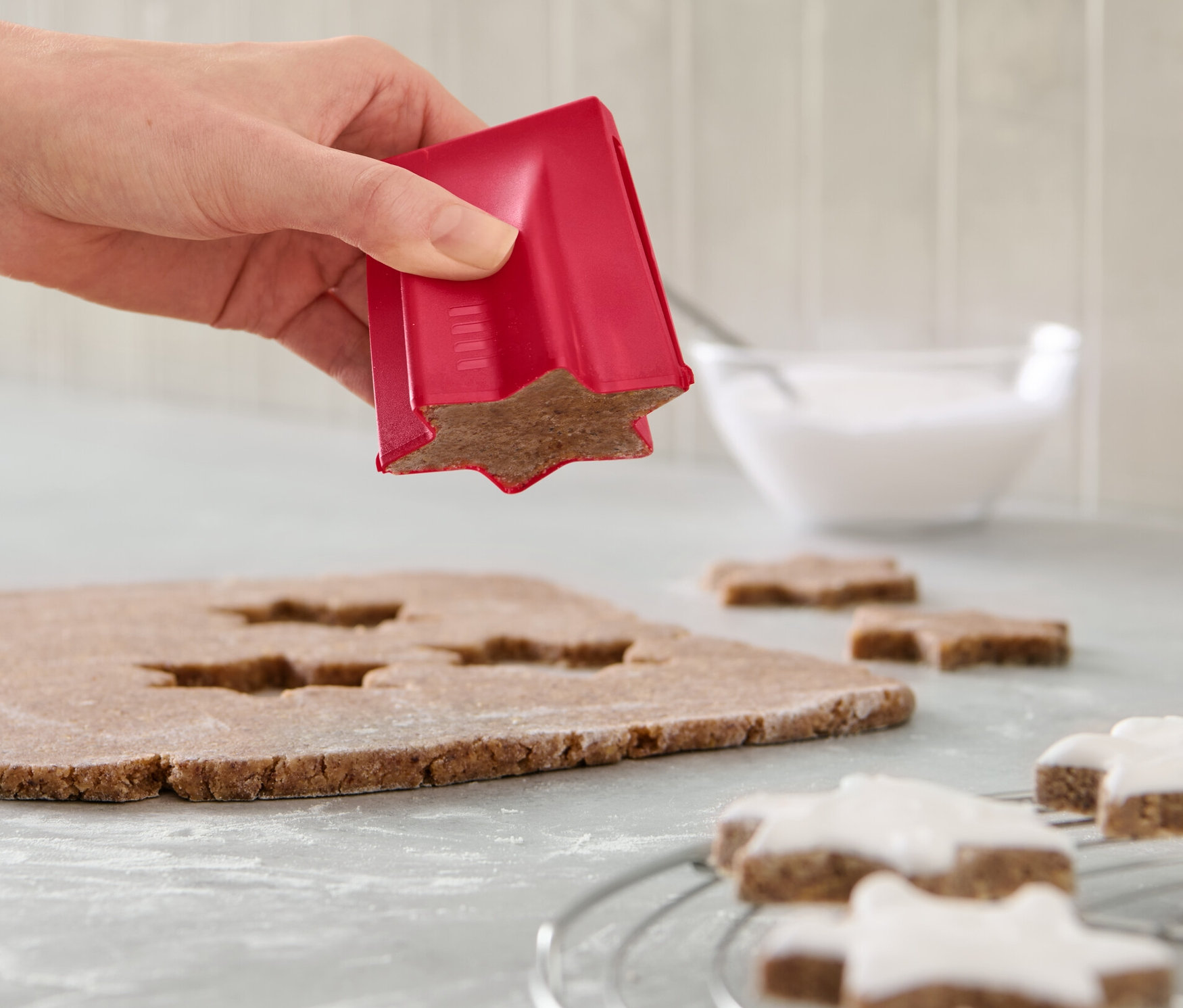 Ręka używa foremki do wykrawania ciastek w kształcie gwiazdy na cieście. Na ladzie leżą ozdobione gwiazdki i miska z lukrem.