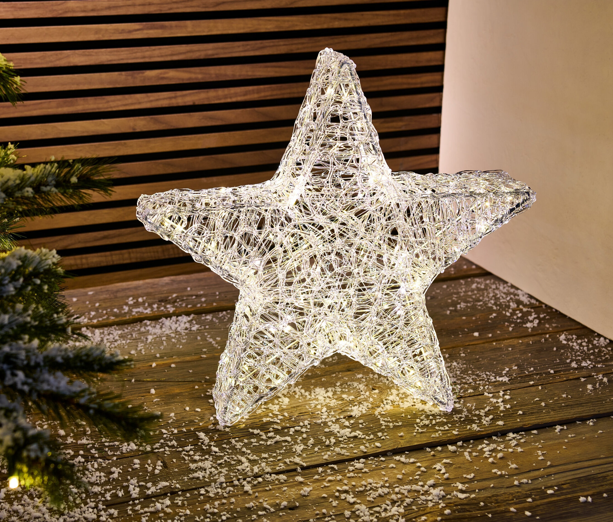 Świecąca dekoracyjna gwiazda LED do użytku zewnętrznego stoi na drewnianej podłodze obok choinki.