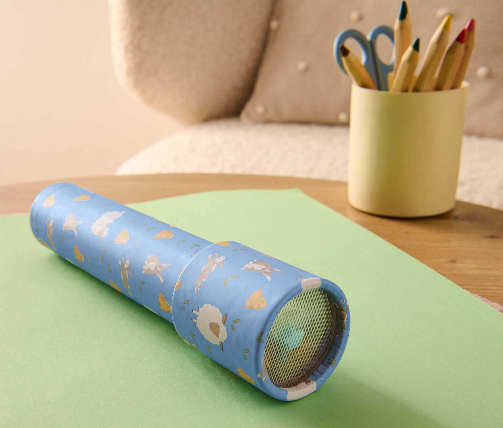 Kalejdoskop leży na zielonym papierze. Obok stoi kubek z kredkami i nożyczkami.