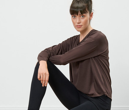 Kobieta siedzi na drewnianym stołku i ma na sobie brązową koszulkę sportową 2 w 1 i czarne legginsy.