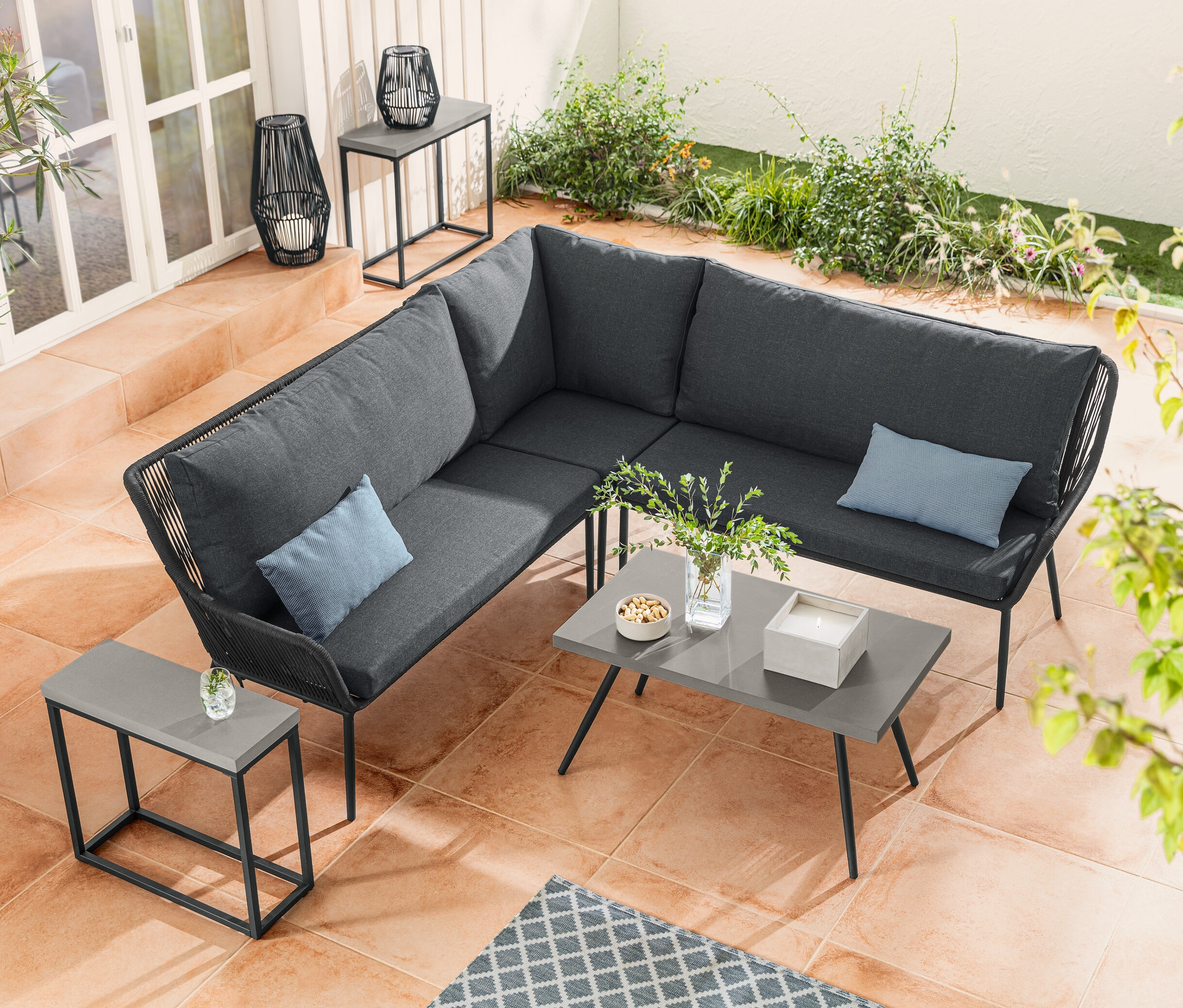 Narożnik wypoczynkowy z plecionką tekstylną stoi obok stolika kawowego z blatem z policementu. Zestaw konsoli z blatem z policementu stoi obok latarni solarnej ze sznurkiem tekstylnym.