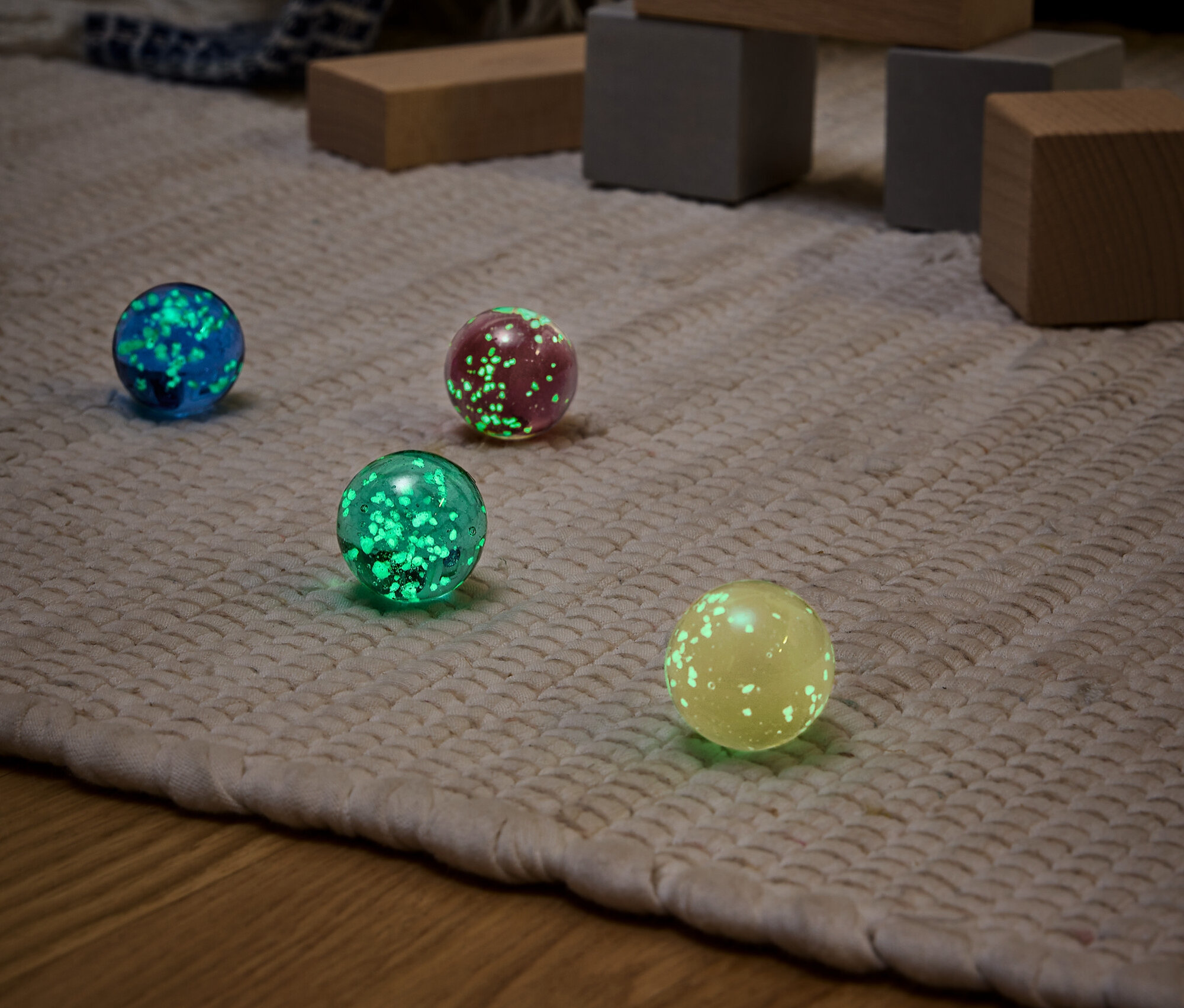 Cztery świecące kule w różnych kolorach (niebieski, czerwony, zielony i żółty) na beżowym dywanie z drewnianymi zabawkami w tle.