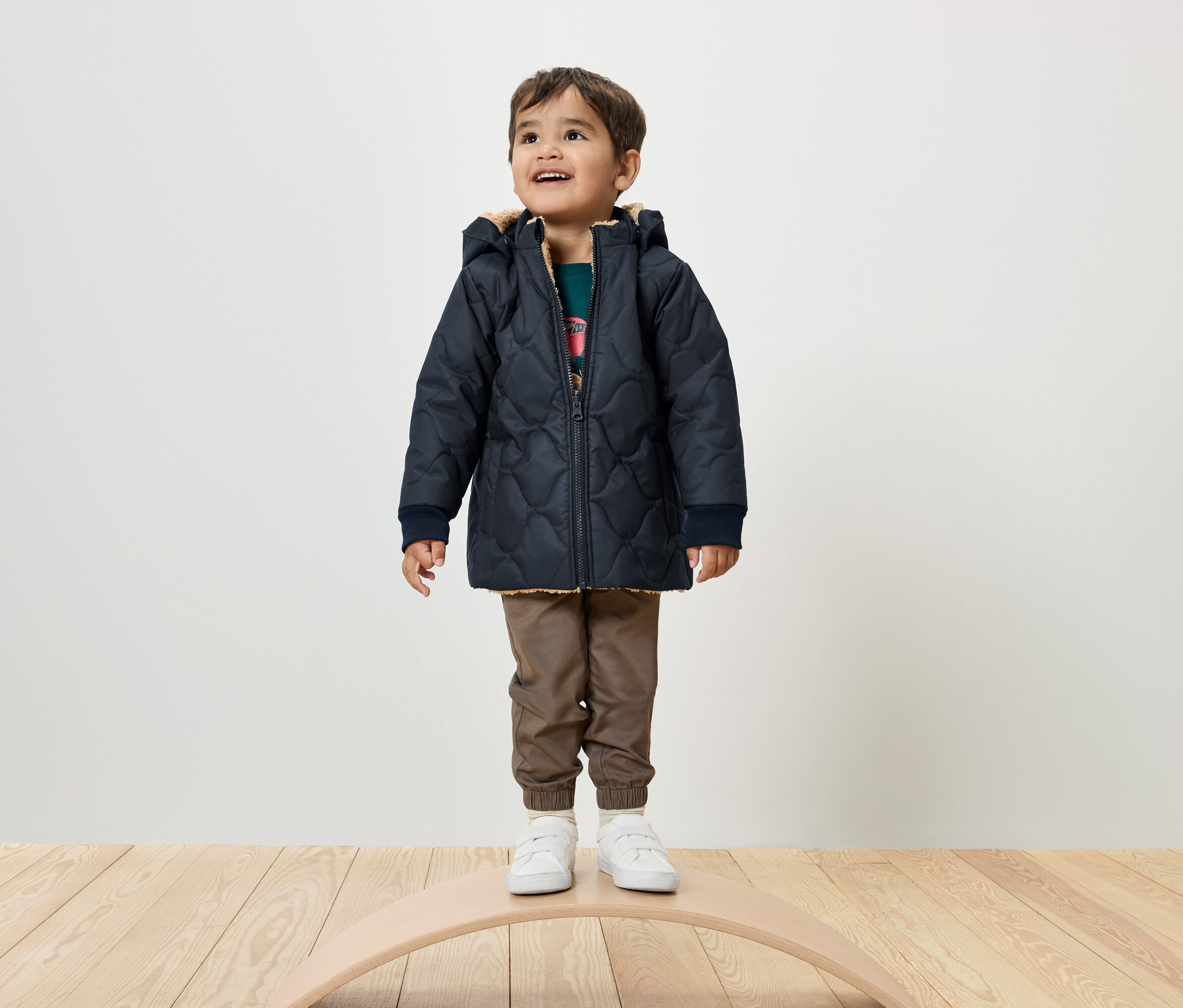 Chłopiec stoi na desce do balansowania, ubrany w granatową kurtkę pikowaną, brązowe spodnie i białe tenisówki.