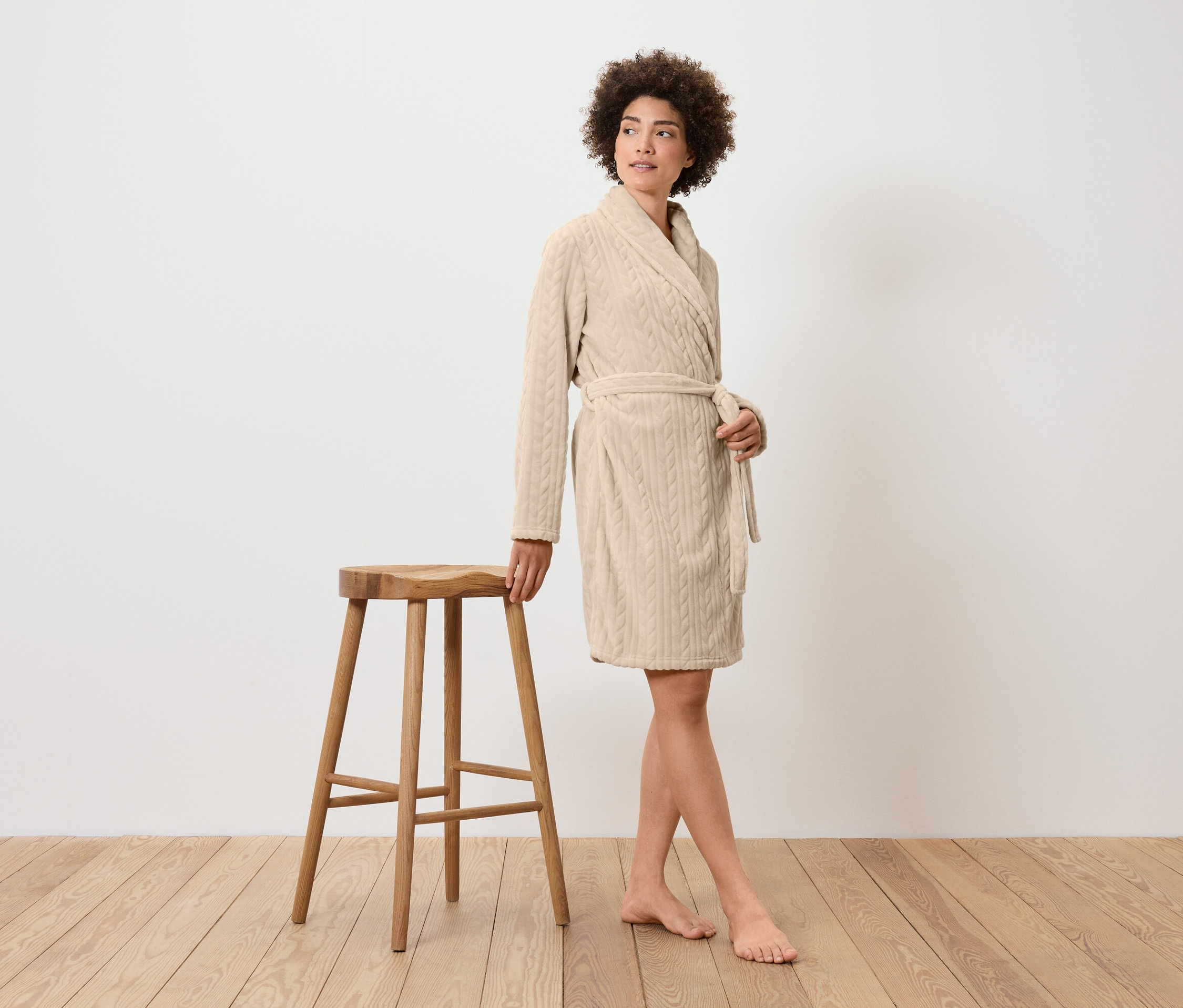 Kobieta w kremowym szlafroku stoi obok drewnianego stołka.