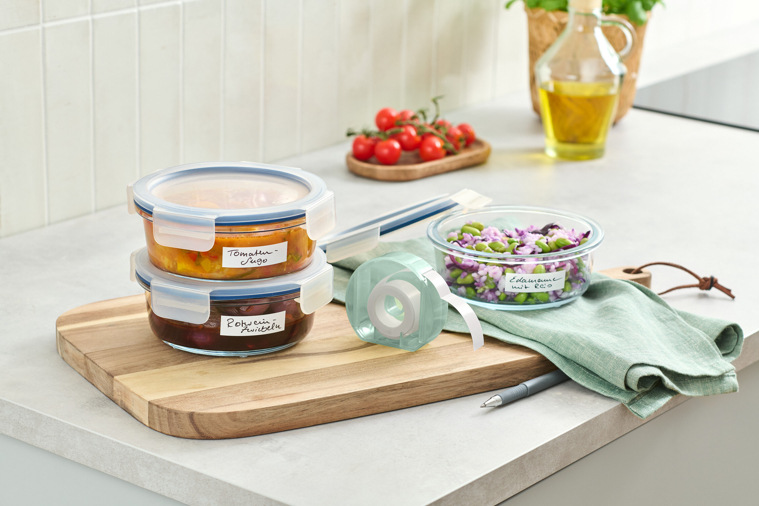 Dwa szklane pojemniki z jedzeniem ułożone na drewnianej desce obok rolki z etykietami.