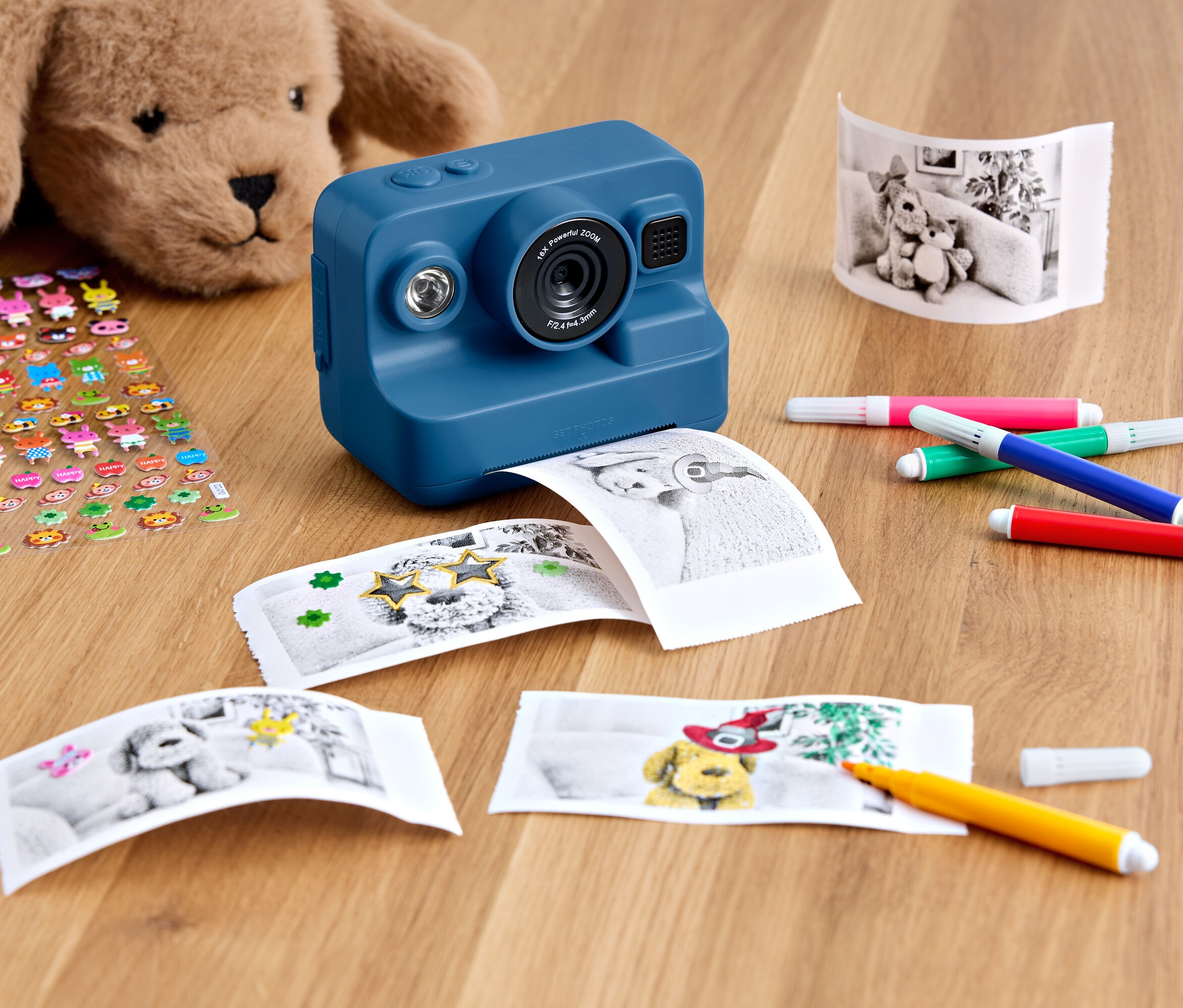 Niebieski aparat natychmiastowy leży na drewnianym stole z pluszowym misiem, arkuszem naklejek, wydrukowanymi zdjęciami i kolorowymi markerami.