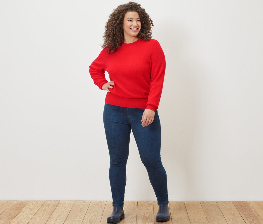 Kobieta pozuje w czerwonym cienkim swetrze i dżinsowych legginsach z wysokim stanem.