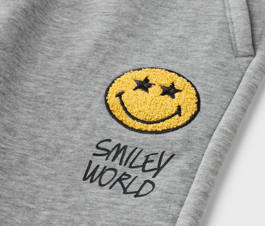 Szczegół szarych joggerów dziecięcych SMILEYWORLD® z naszywką z żółtą buźką.
