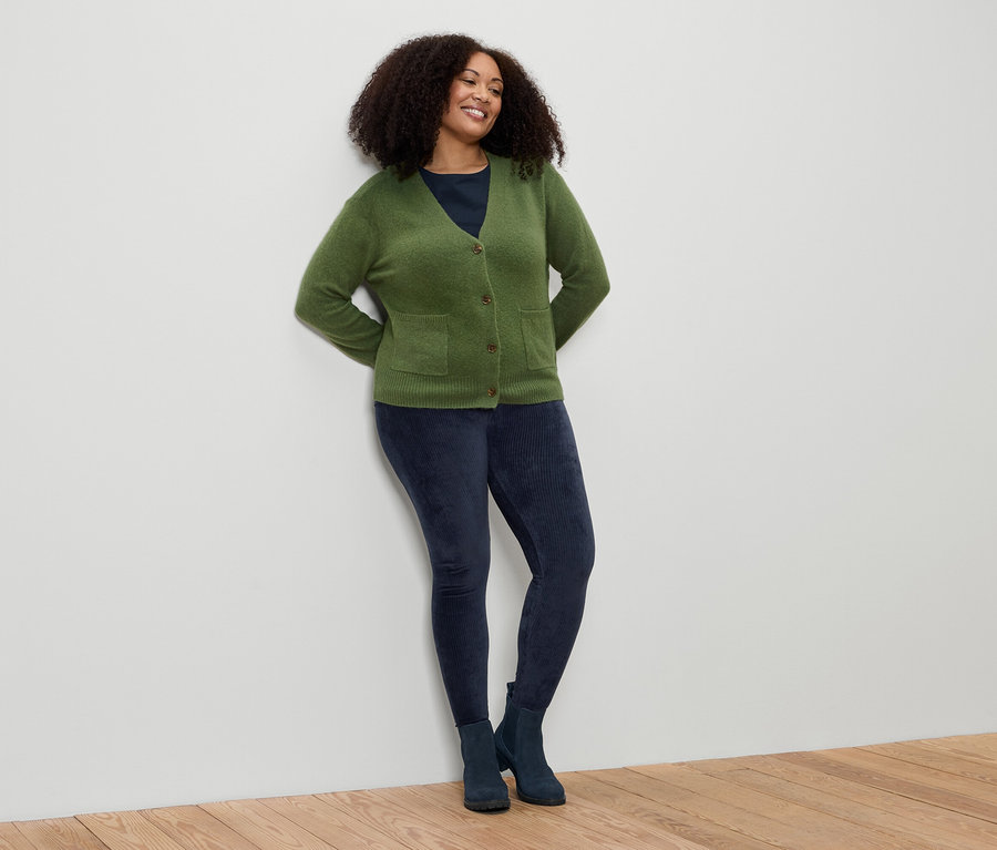 Kobieta z kręconymi włosami opiera się o ścianę. Ma na sobie zielony kardigan, niebieskie legginsy i buty.