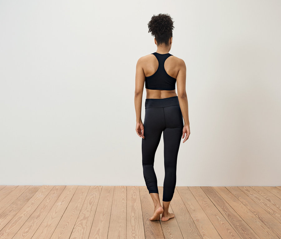 Kobieta w czarnym stroju sportowym stoi tyłem do kamery na drewnianej podłodze.