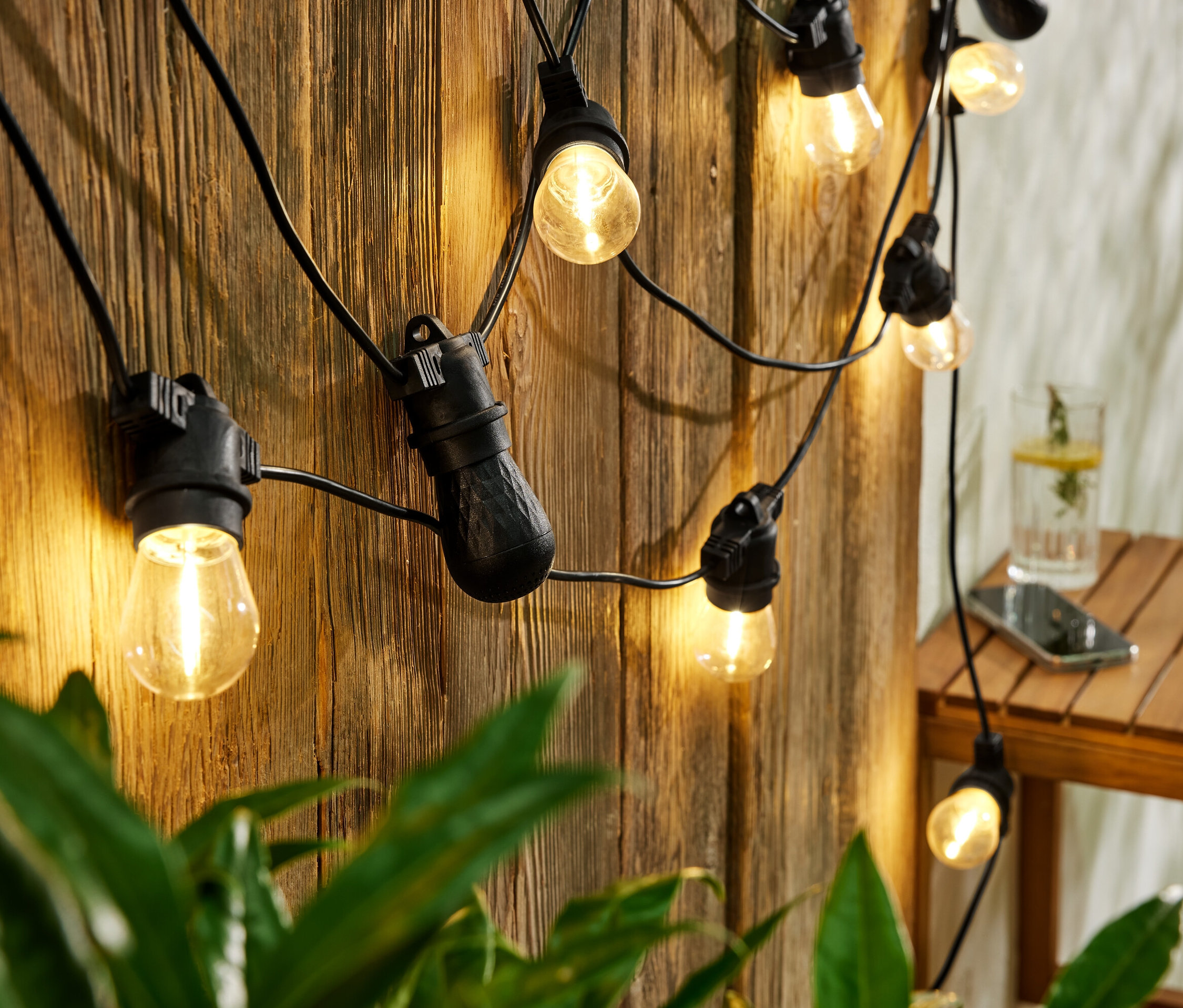 Na drewnianej ścianie wisi łańcuch świetlny LED z głośnikami Bluetooth do użytku na zewnątrz. Na stole szklanka z napojem i telefon.