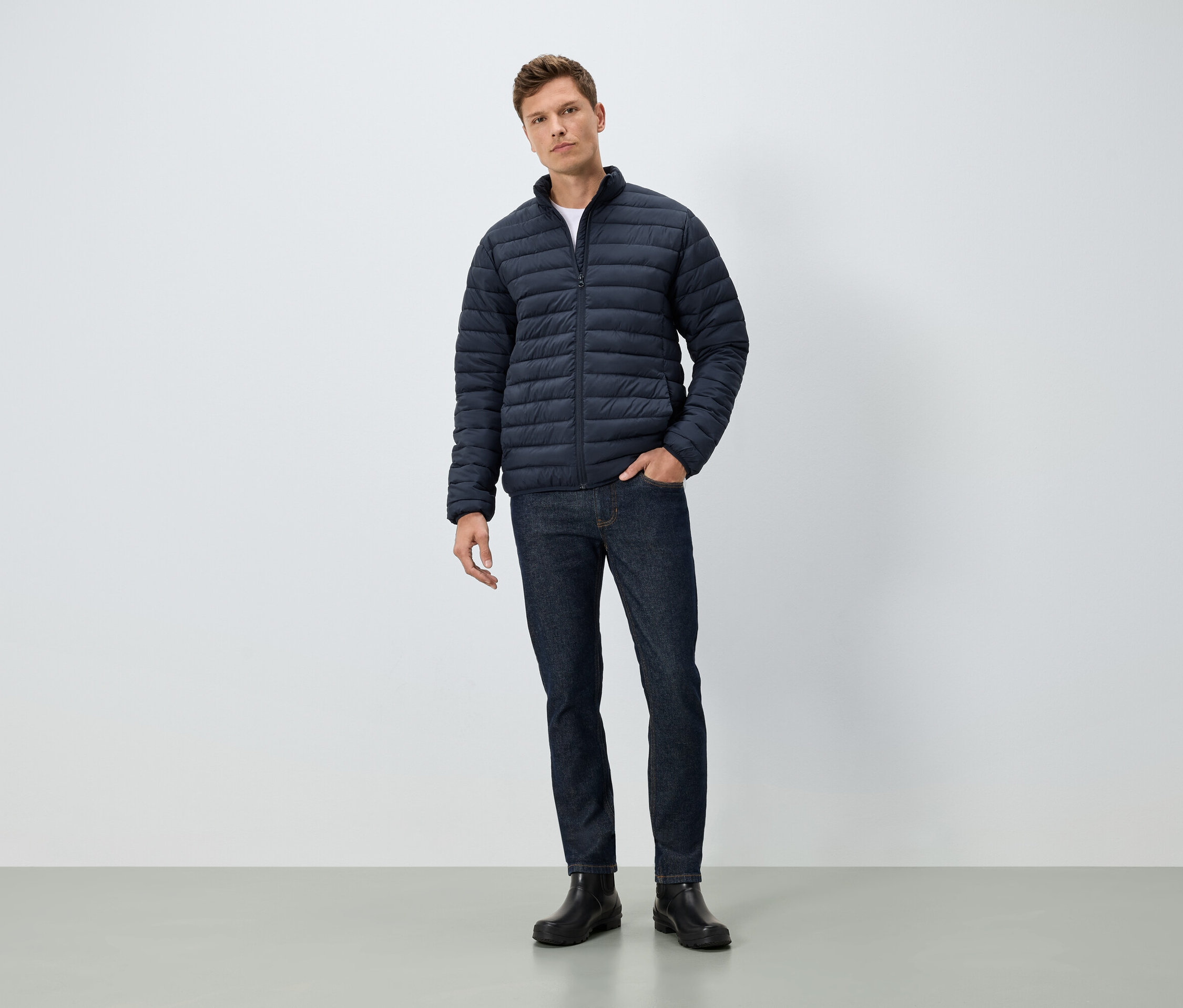 Mężczyzna stoi z ręką w kieszeni. Ma na sobie niebieską kurtkę, dżinsy i czarne buty.