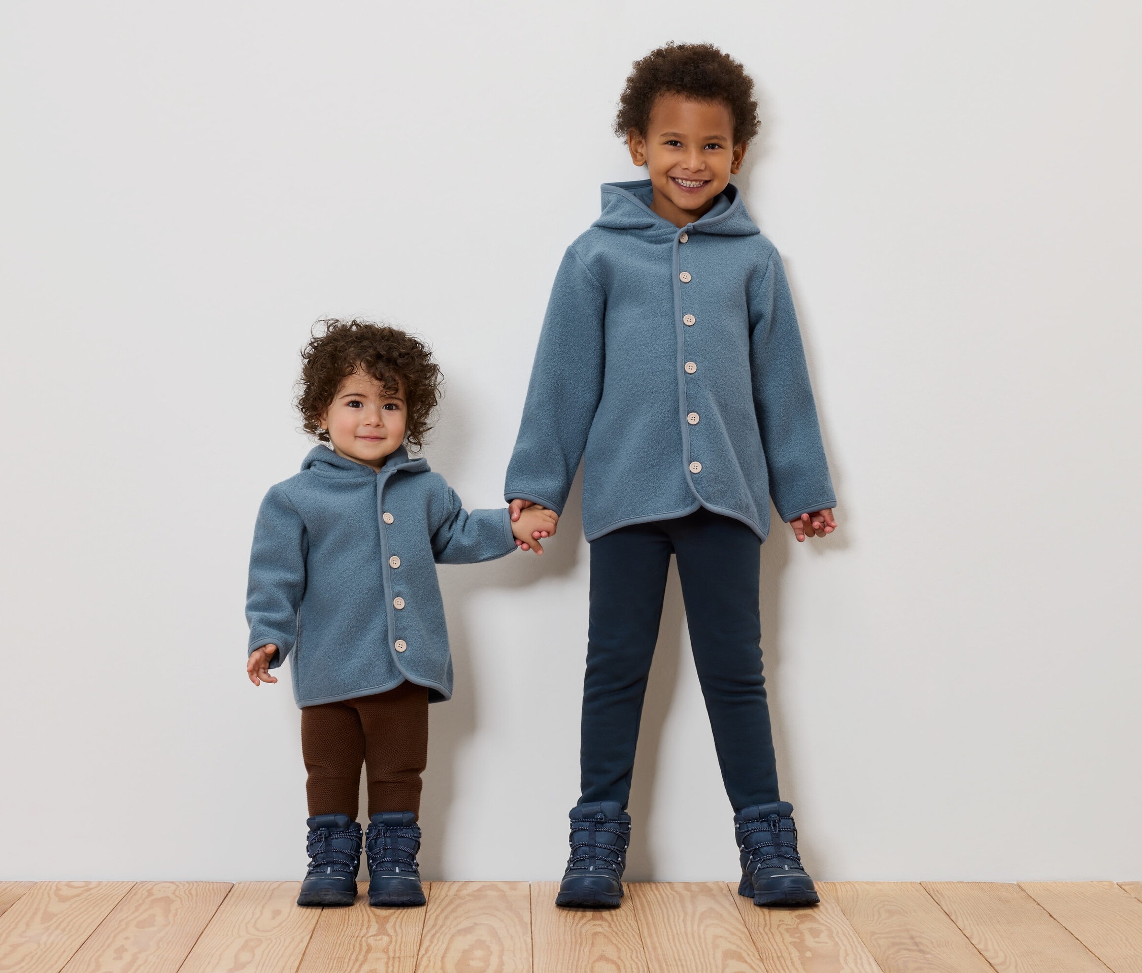 Dwoje dzieci trzyma się za ręce. Oboje mają na sobie niebieskie płaszcze i buty.