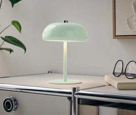 Zielona lampa stoi na białym stoliku nocnym obok rośliny i okularów.