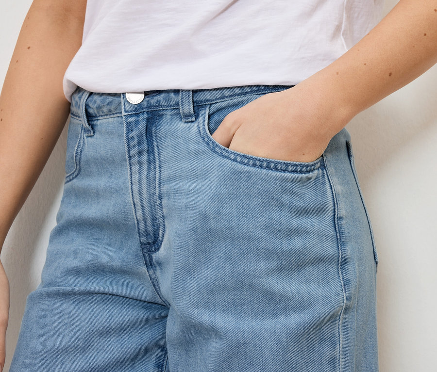 Zbliżenie kobiety w jasno niebieskich jeansach i białym T-shircie, trzymającej rękę w kieszeni.