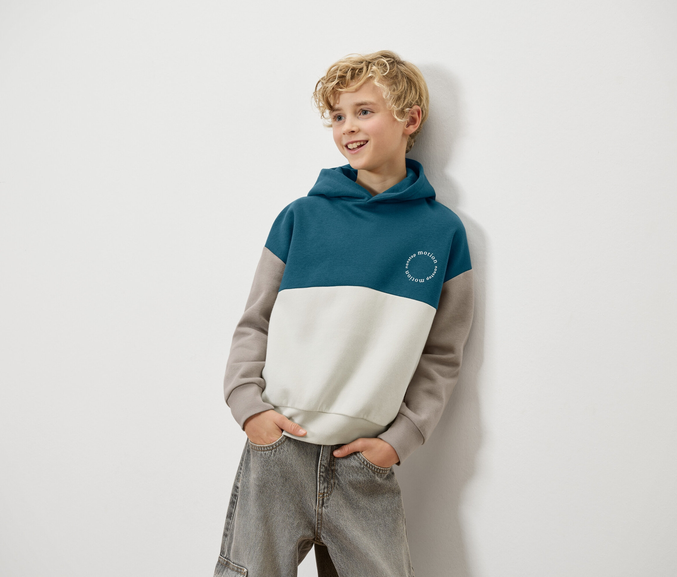 Blond chłopiec opiera się o ścianę, ubrany w niebiesko-biało-brązową bluzę z kapturem i szare dżinsy.