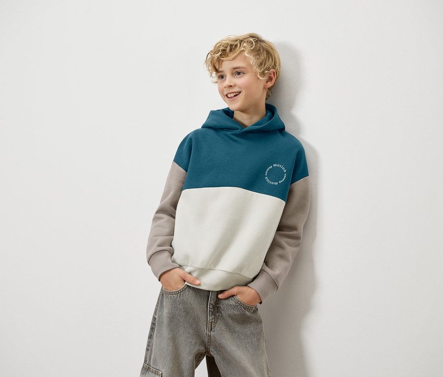 Blond chłopiec opiera się o ścianę, ubrany w niebiesko-biało-brązową bluzę z kapturem i szare dżinsy.