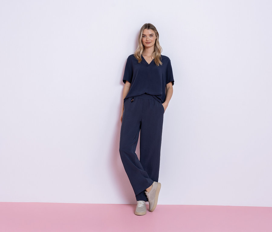 Kobieta stoi na różowo-białym tle, ubrana w granatowy strój składający się z T-shirtu i spodni oraz szare trampki.