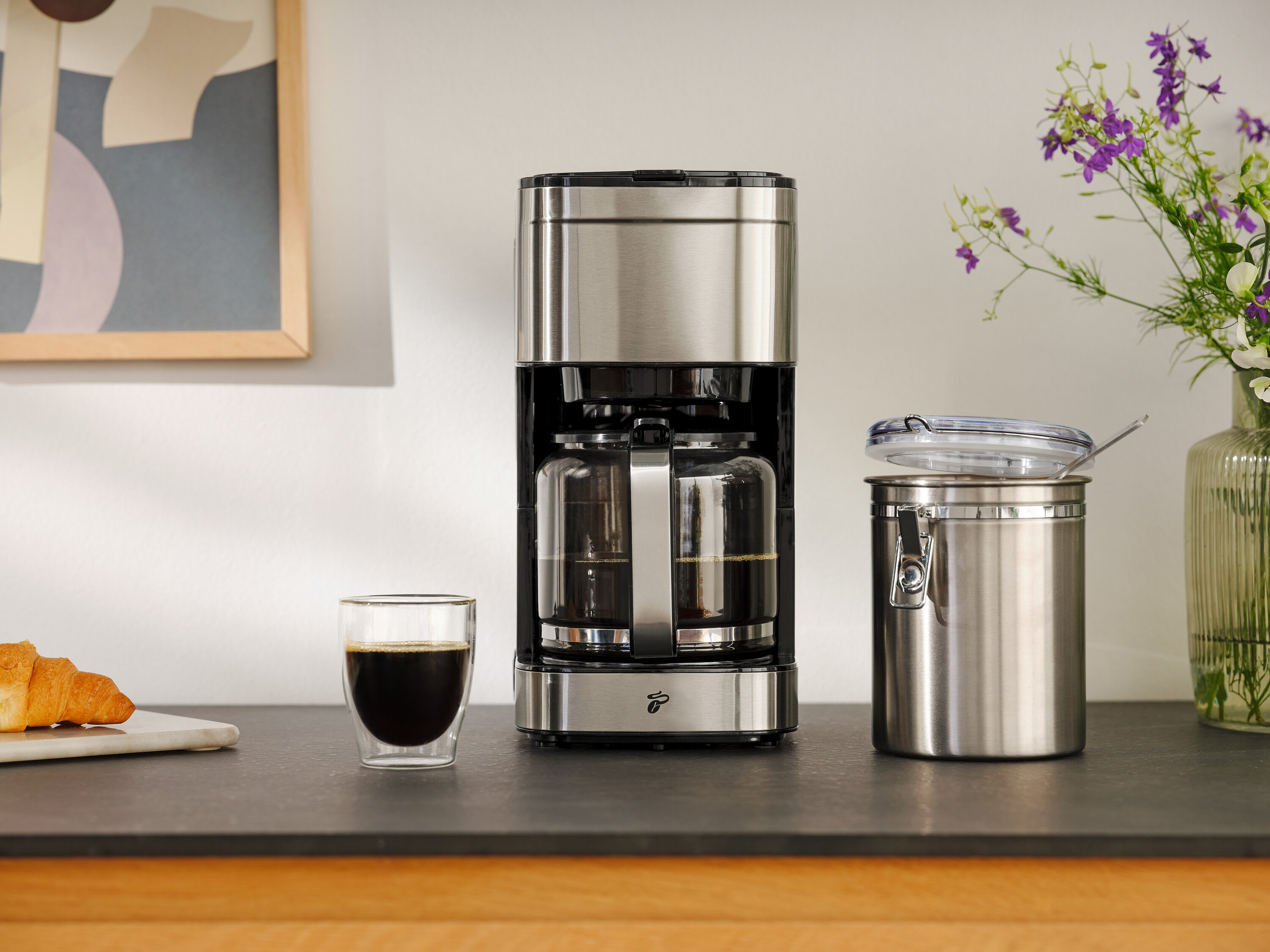 Ekspres do kawy obok szklanki kawy, rogalika i pojemnika do przechowywania.