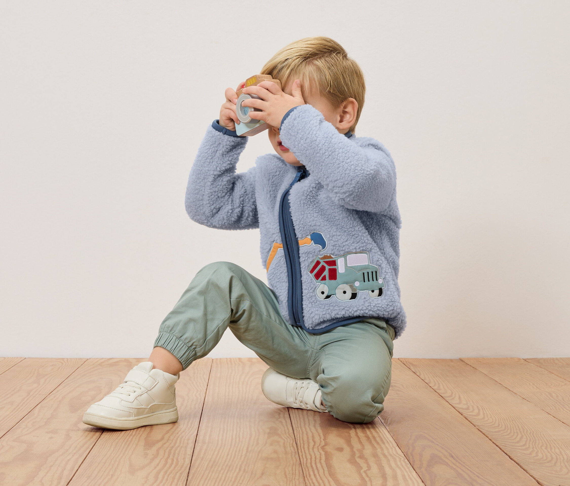 Blondwłosy chłopiec klęczy na drewnianej podłodze i trzyma przed twarzą zabawkę-aparat. Ma na sobie niebieską bluzę polarową z aplikacją ciężarówki i zielone spodnie.