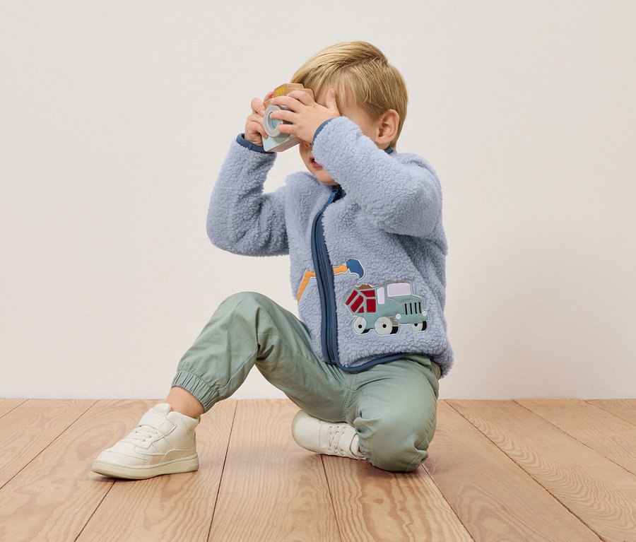 Blondwłosy chłopiec klęczy na drewnianej podłodze i trzyma przed twarzą zabawkę-aparat. Ma na sobie niebieską bluzę polarową z aplikacją ciężarówki i zielone spodnie.