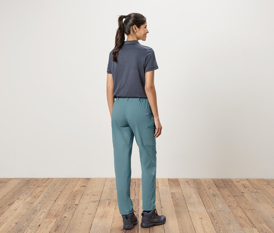 Widok z tyłu kobiety w kucyku, ubranej w granatową koszulkę polo, zielone spodnie i buty trekkingowe na tle białej ściany i drewnianej podłogi.