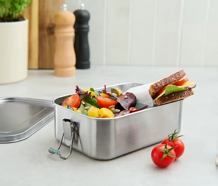 Metalowy pojemnik na lunch ze sałatką i kanapką, obok leżą dwa pomidory.