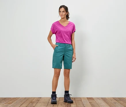 Kobieta stoi z ręką w kieszeni, ubrana w różowy t-shirt, zielone szorty i buty trekkingowe.
