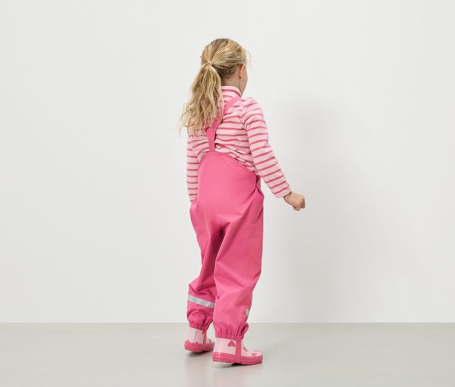 Widok z tyłu na dziewczynę o blond włosach, ubraną w różowo-biały sweter w paski, różowe spodnie przeciwdeszczowe na szelkach i różowe kalosze w serduszka.