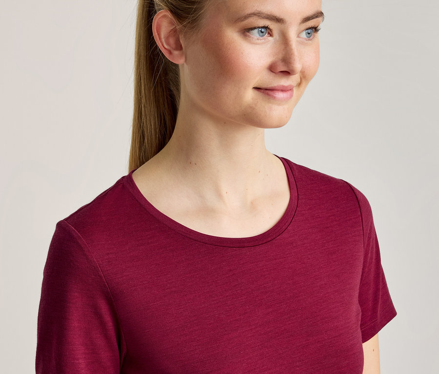 Młoda kobieta z blond włosami spiętymi w kucyk, ubrana w bordowy T-shirt z okrągłym dekoltem, uśmiecha się i patrzy w prawo.