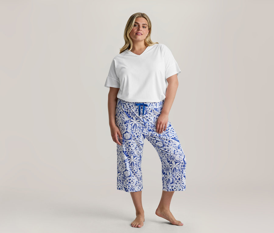 Kobieta stoi, ubrana w biały T-shirt z dekoltem w serek i niebiesko-białe wzorzyste spodnie od 3/4-Pyjama-Set z niebieskim sznurkiem, boso.
