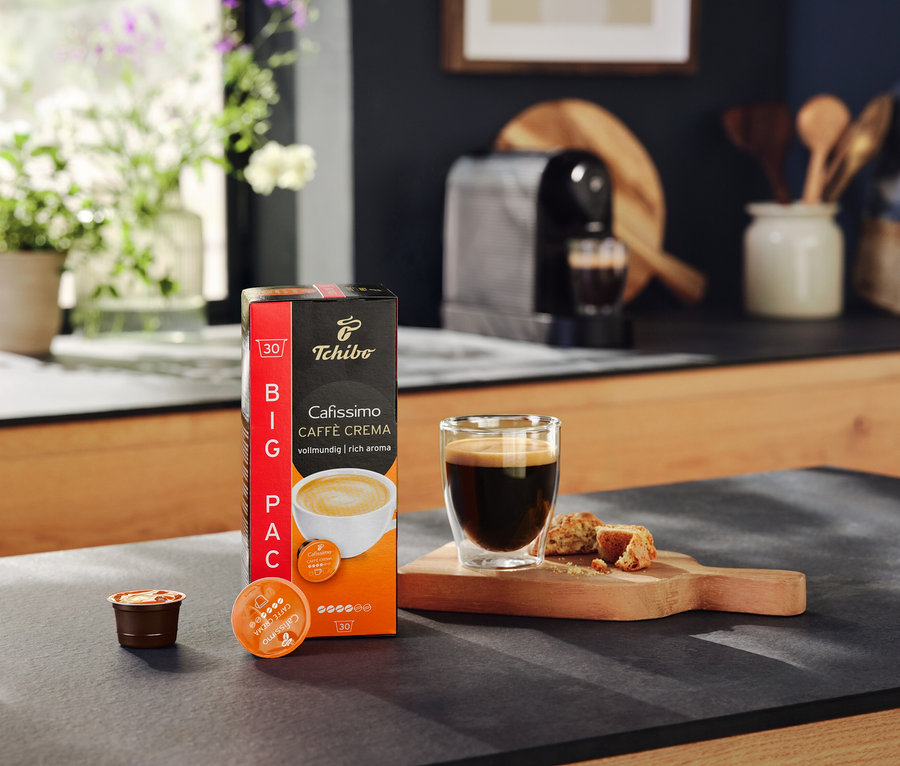 Opakowanie kapsułek Cafissimo Caffe Crema, szklanka kawy i ciasteczka na stole. W tle ekspres do kawy Cafissimo pure+ black.