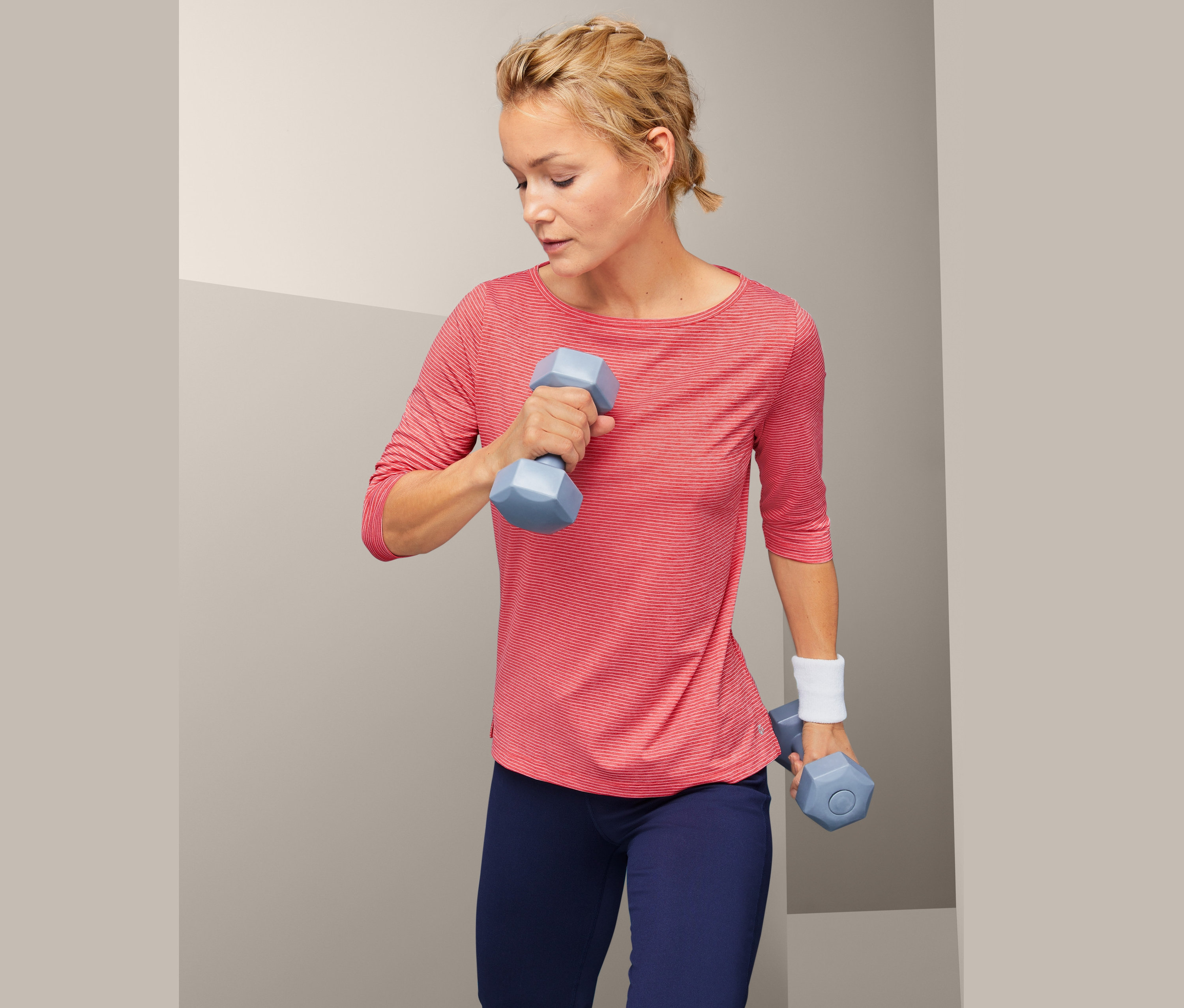 Kobieta ćwiczy z niebieskimi hantlami, ubrana w koszulkę funkcyjną.