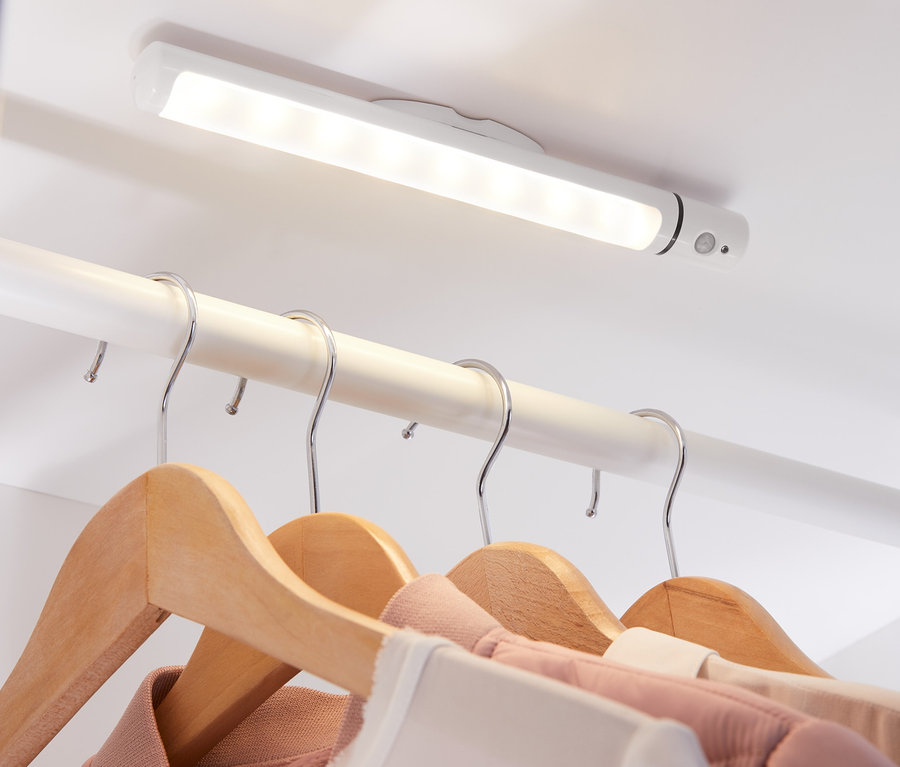 Wielofunkcyjna lampa LED przymocowana do sufitu szafy oświetla ubrania wiszące na drewnianych wieszakach.