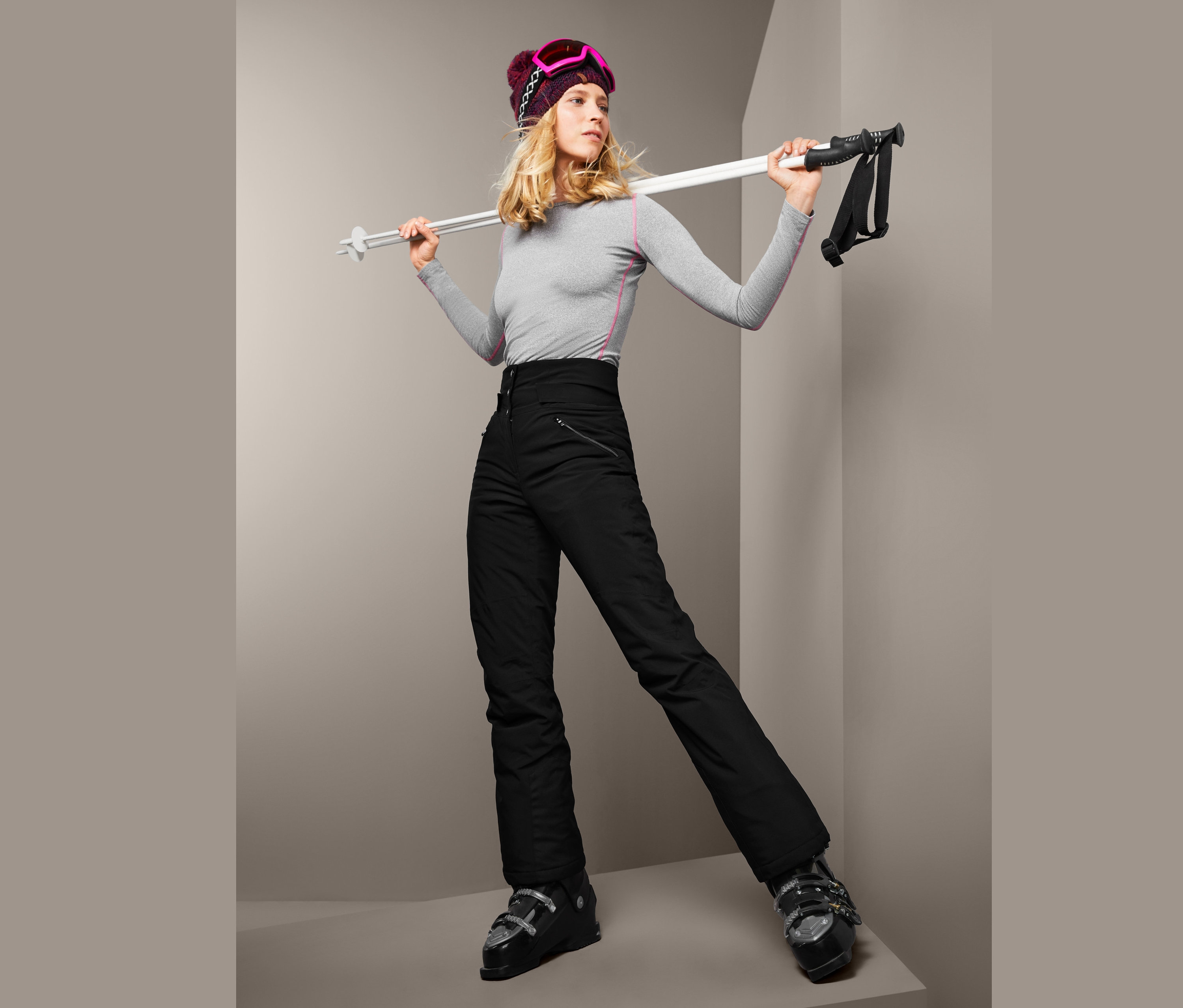 Blondynka w szarej koszulce i czarnych damskich spodniach narciarskich softshell z wysokim stanem trzyma kijki narciarskie.
