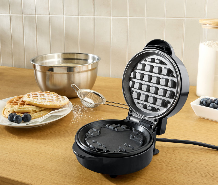 Otwarta, czarna gofrownica na drewnianym blacie obok talerza gofrów posypanych cukrem pudrem i borówkami.