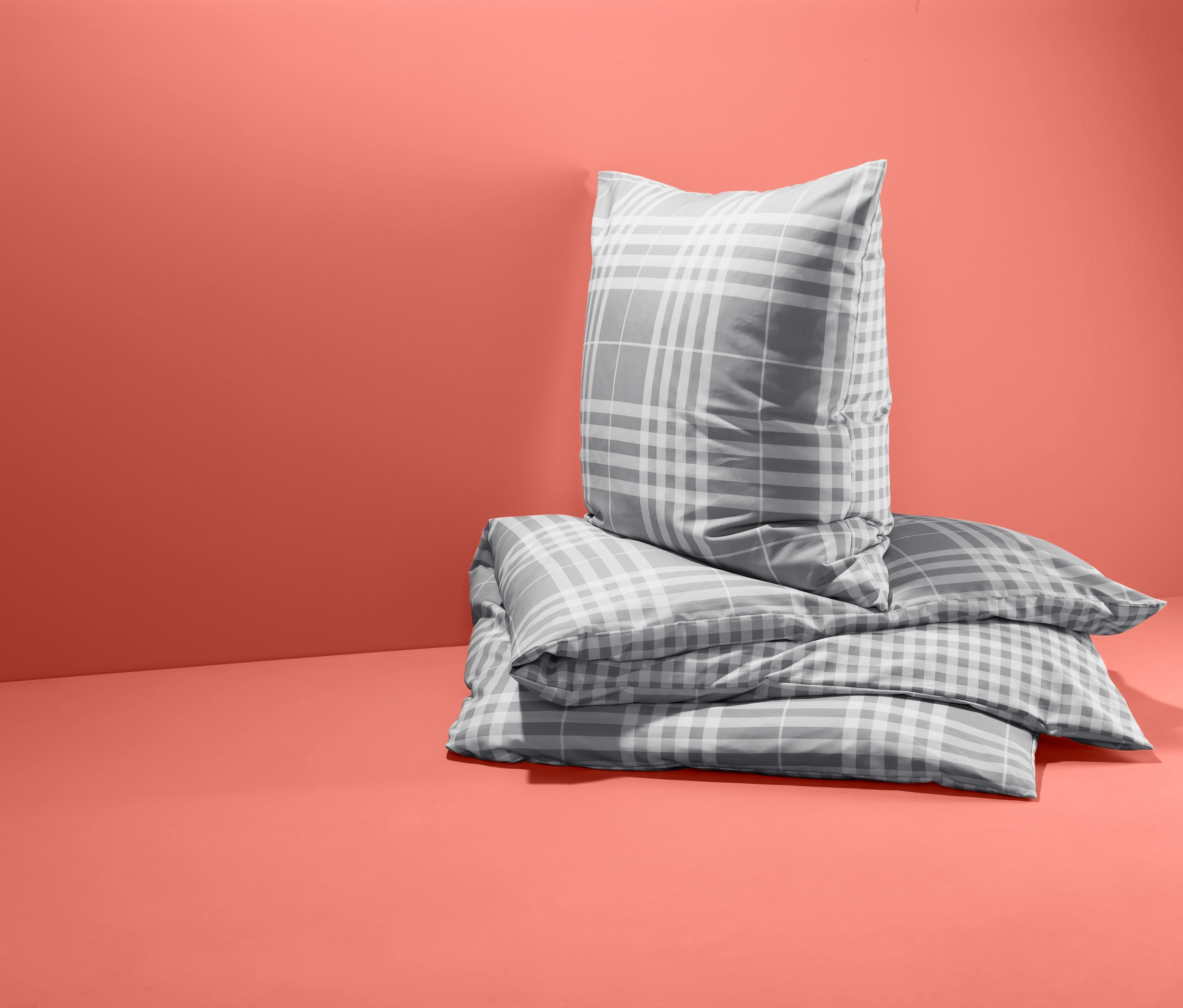 Na koralowym tle leżą na sobie trzy zestawy dwustronnej pościeli w kratę ze wzmocnionej bawełny w standardowym rozmiarze.