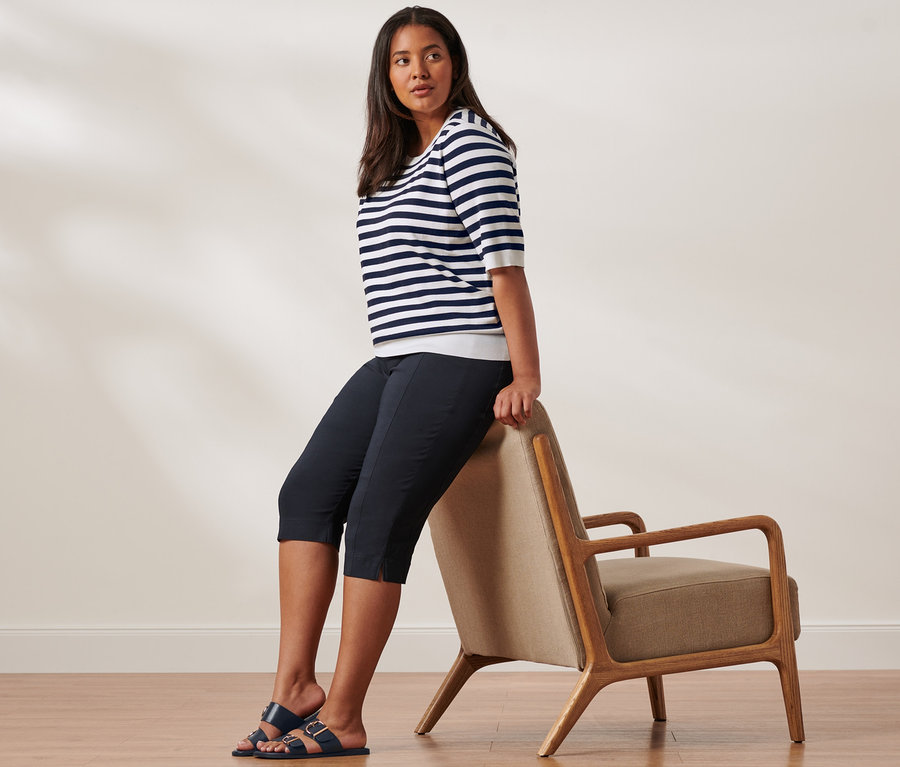 Kobieta siedzi na oparciu krzesła, ubrana w niebiesko-biały sweter z delikatnej dzianiny, spodnie stretchowe o długości 3/4 i sandały ze sprzączką.