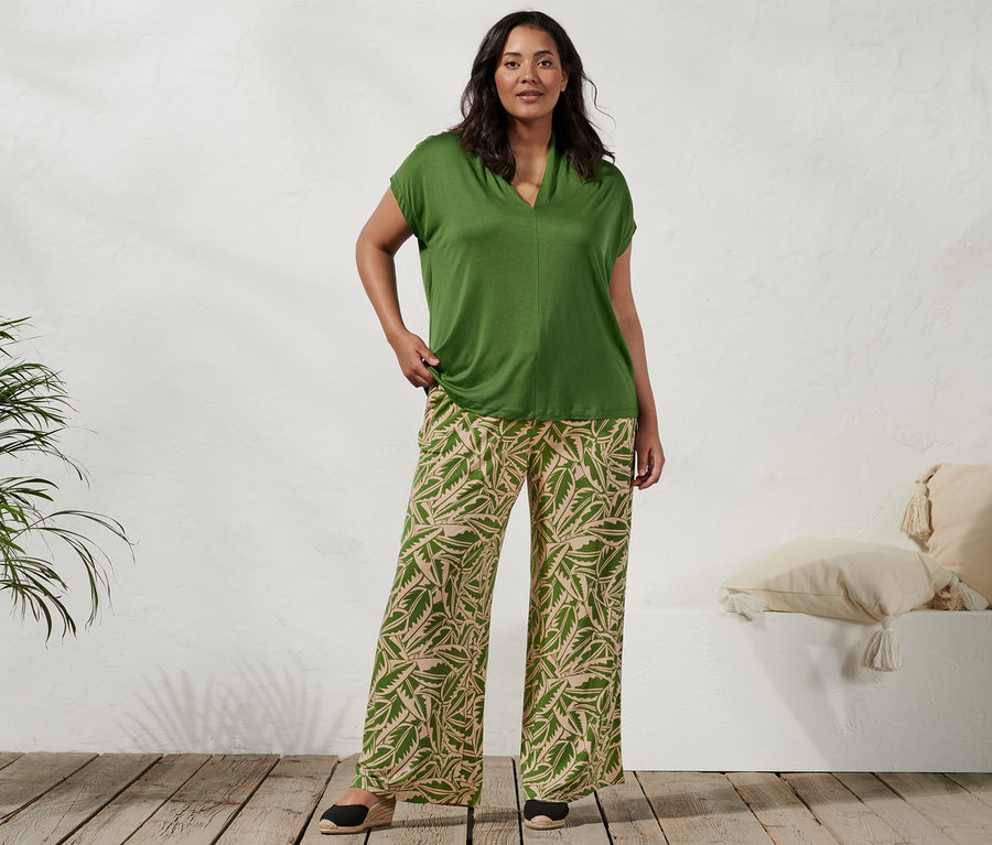 Kobieta stoi i ma na sobie zieloną bluzkę ze stójką, zielono-beżowe spodnie palazzo z nadrukiem i espadryle na koturnie.