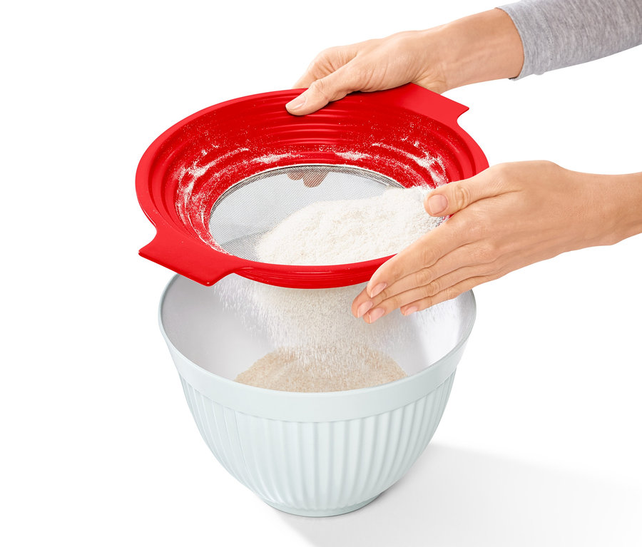 Kobieta przesiewa mąkę przez czerwone sitko do przesiewania mąki i przecierania 2 w 1 do białej miski.