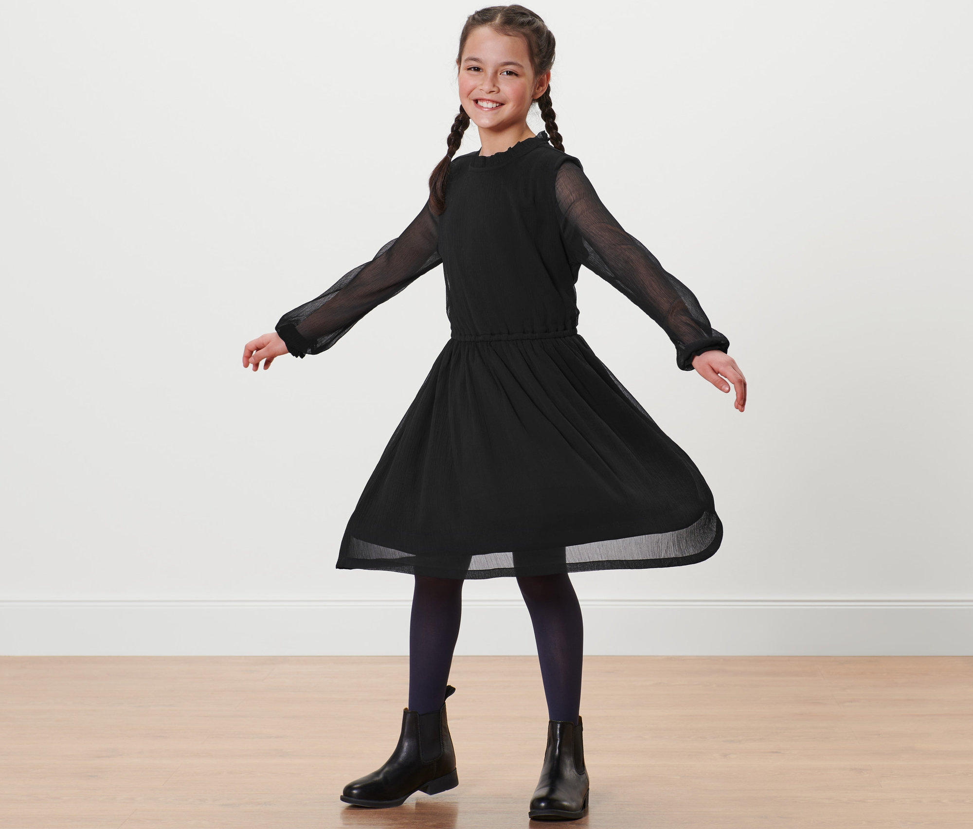 Uśmiechnięta dziewczyna w warkoczach stoi w czarnej sukience szyfonowej.