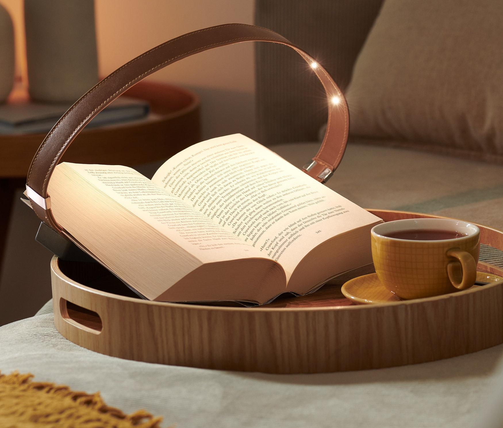 Otwarta książka leży na drewnianej tacy z lampką do czytania z magnetycznym klipsem i filiżanką herbaty.