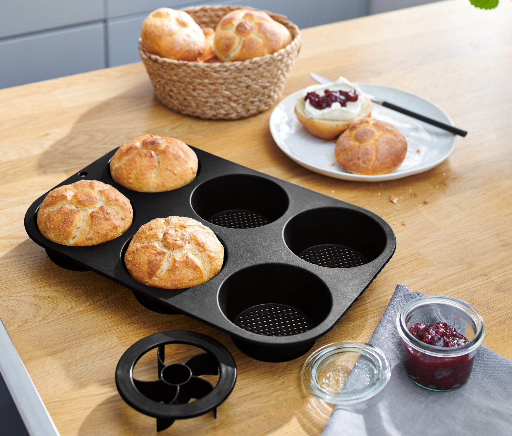 Cztery bułki w czarnej, perforowanej formie do pieczenia bułek na drewnianym stole.