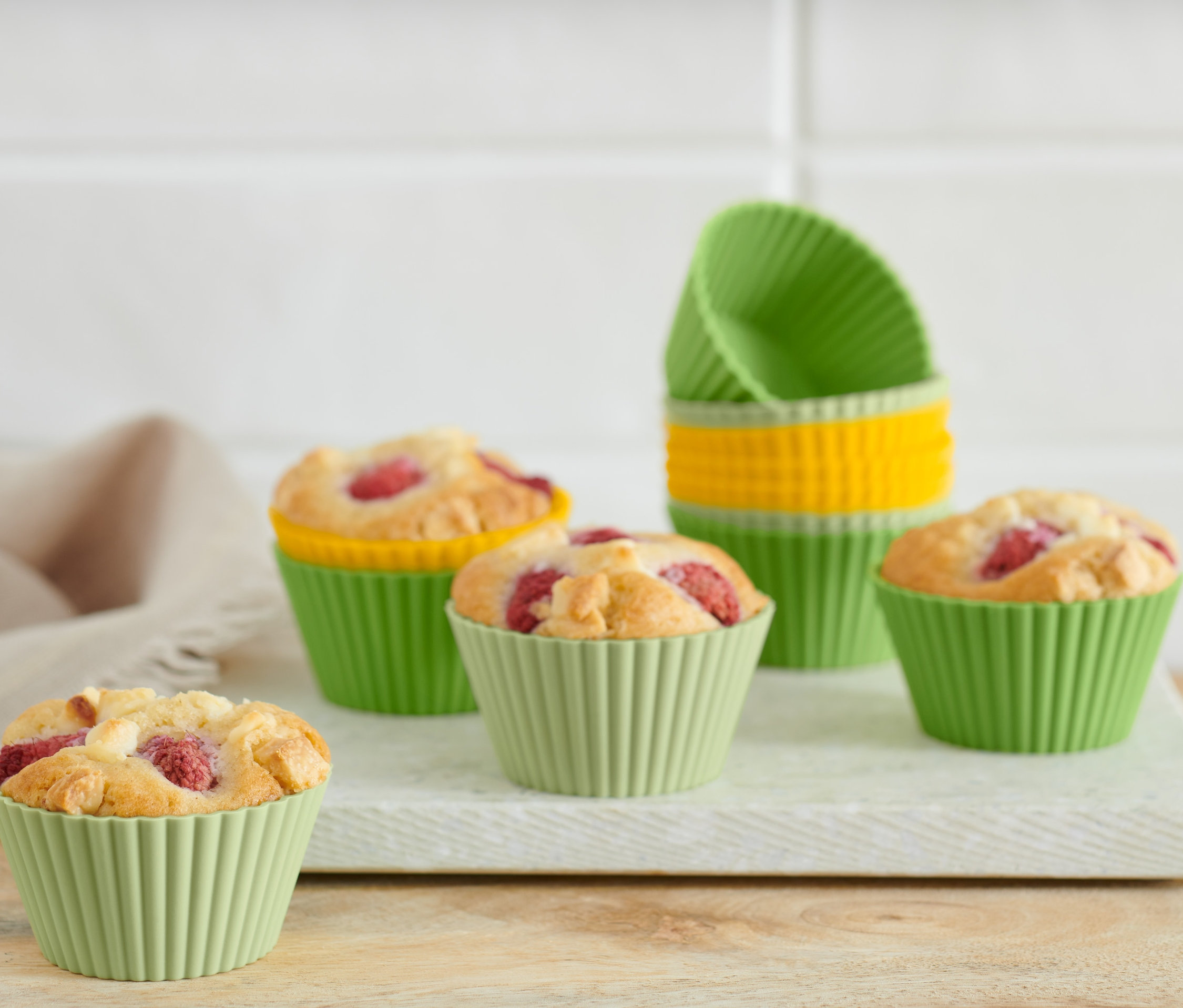 Pięć babeczek w zielonych foremkach na białej desce. W tle widoczne foremki do muffinów, 12 sztuk ułożone w stos.