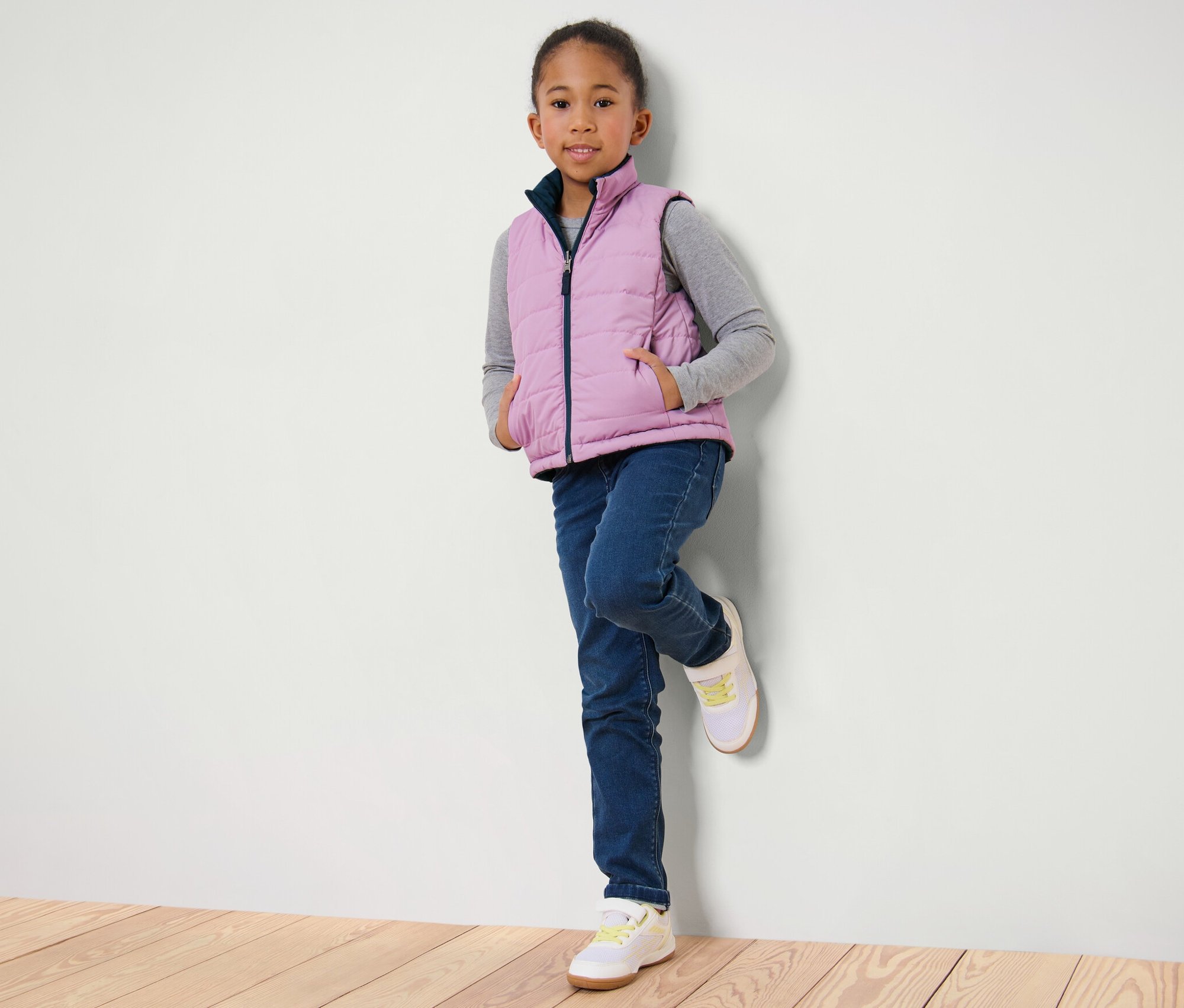 Dziewczyna oparta o ścianę ma na sobie dziecięcą kamizelkę pikowaną, dżinsy i tenisówki.