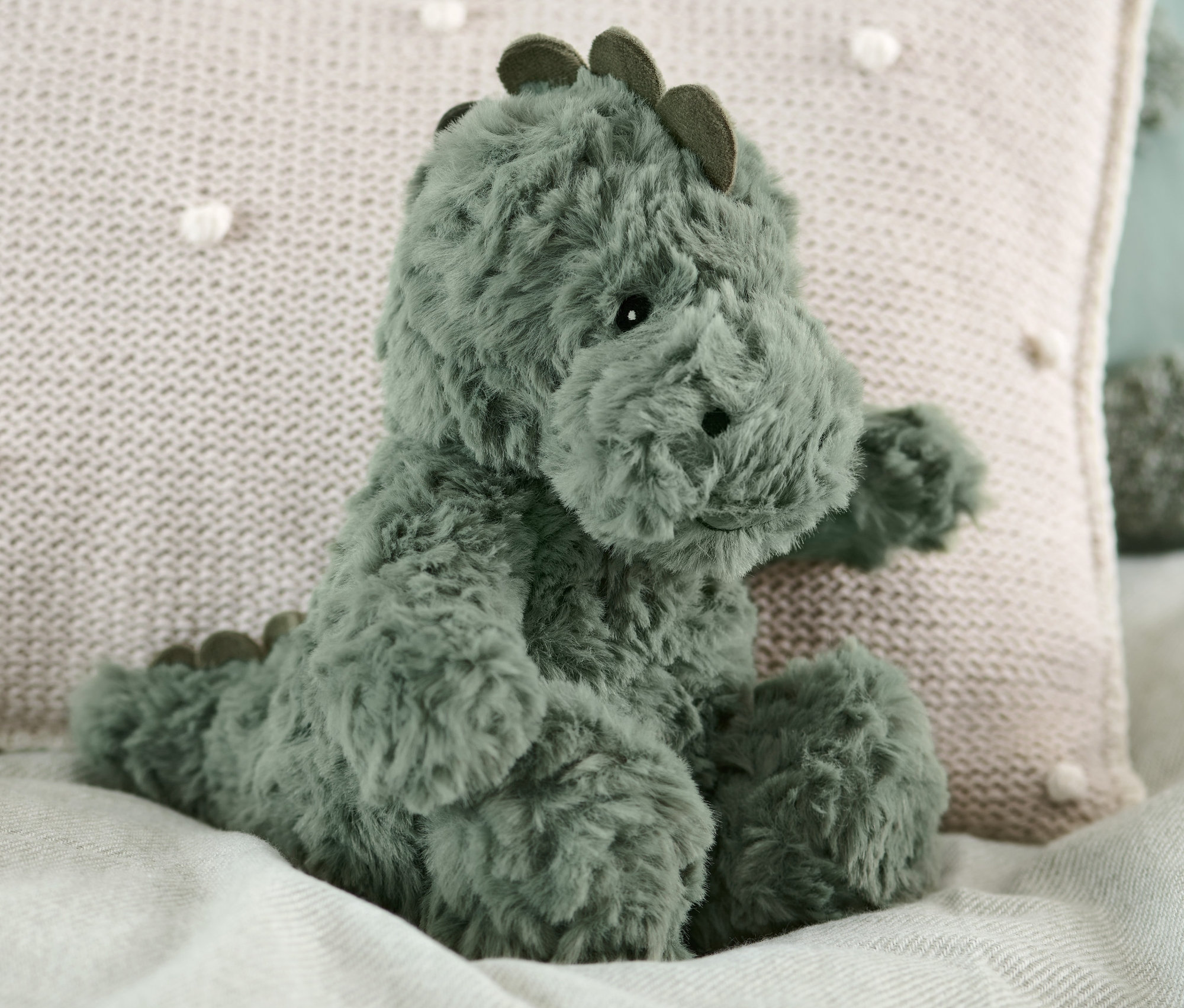 Pluszowy dinozaur siedzi na łóżku obok beżowej poduszki.