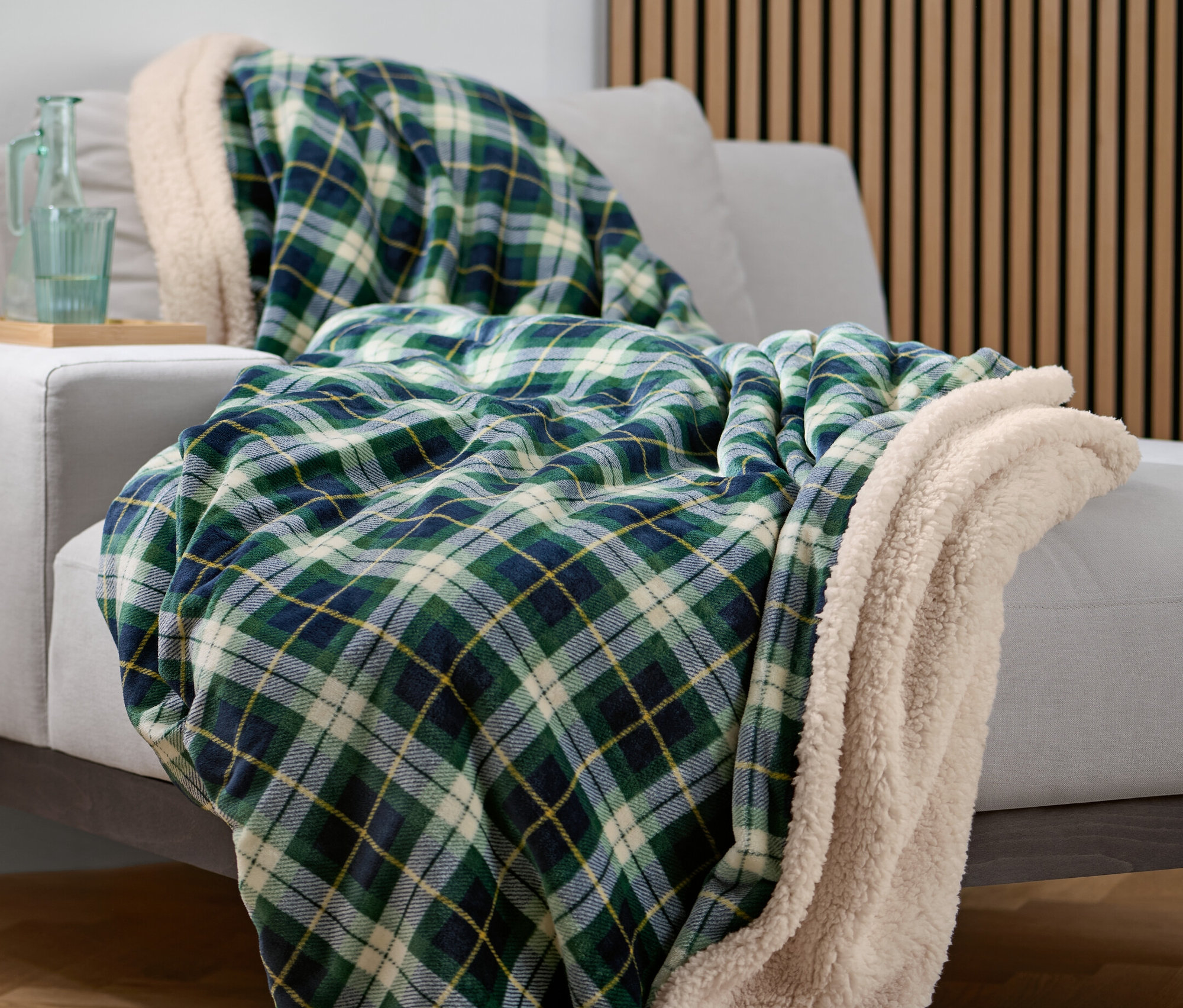 Zielony koc w kratę leży na szarej sofie. Na stoliku bocznym stoi szklana karafka i szklanka.