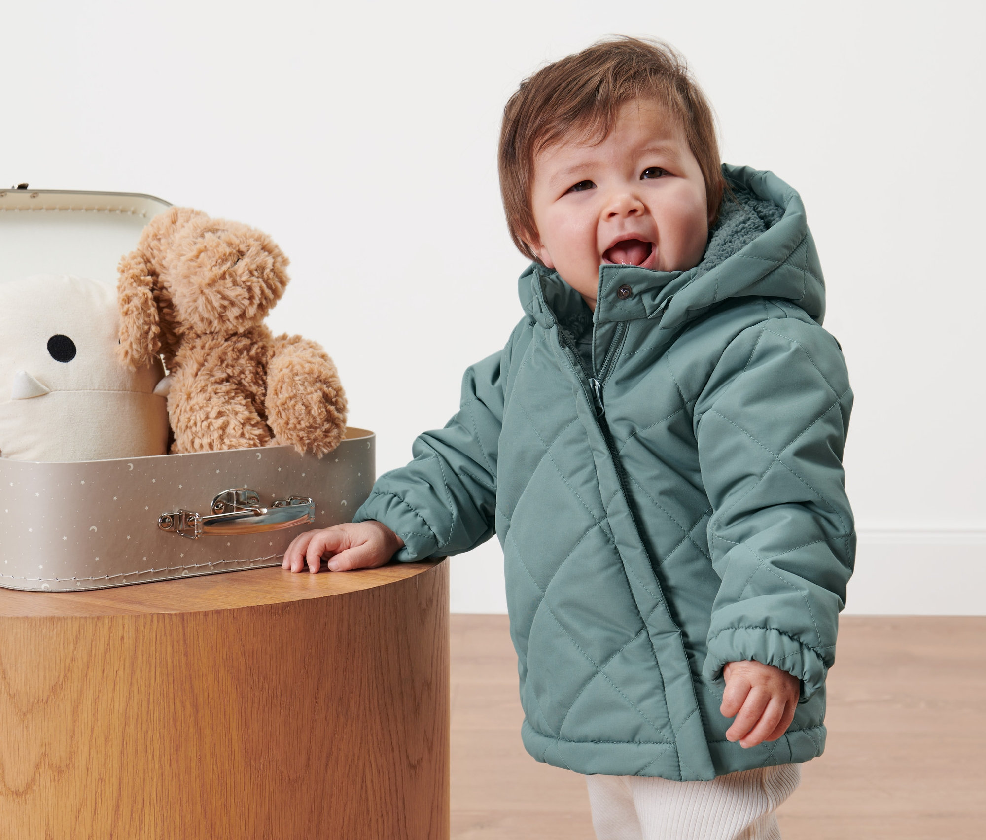 Małe dziecko w pikowanej kurtce stoi obok walizki z zabawkami.